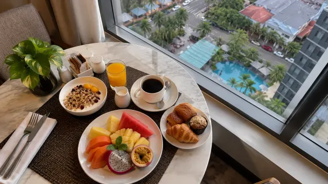 A breakfast table by a window with tropical fruit and coffee of a 4-star city hotel — AP Premier Batam, a 4-star city hotel in Harbour Bay, Batam